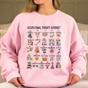 May include: Pink sweatshirt with the text "Occupational Therapy Alphabet" and a grid of 26 illustrated icons representing different aspects of occupational therapy, each icon is labeled with a letter of the alphabet.