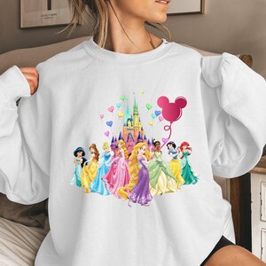 May include: White sweatshirt featuring a colourful graphic of Disney princesses and Cinderella's castle. The graphic also includes a pink Mickey Mouse balloon with hearts.