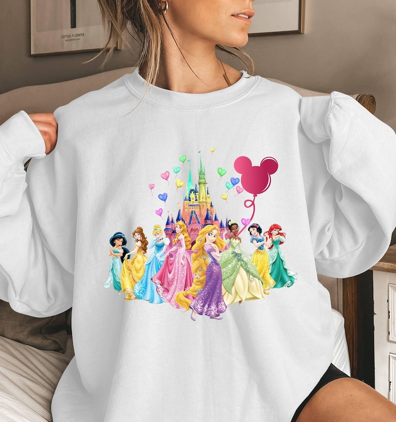 May include: White sweatshirt featuring a colourful graphic of Disney princesses and Cinderella's castle. The graphic also includes a pink Mickey Mouse balloon with hearts.
