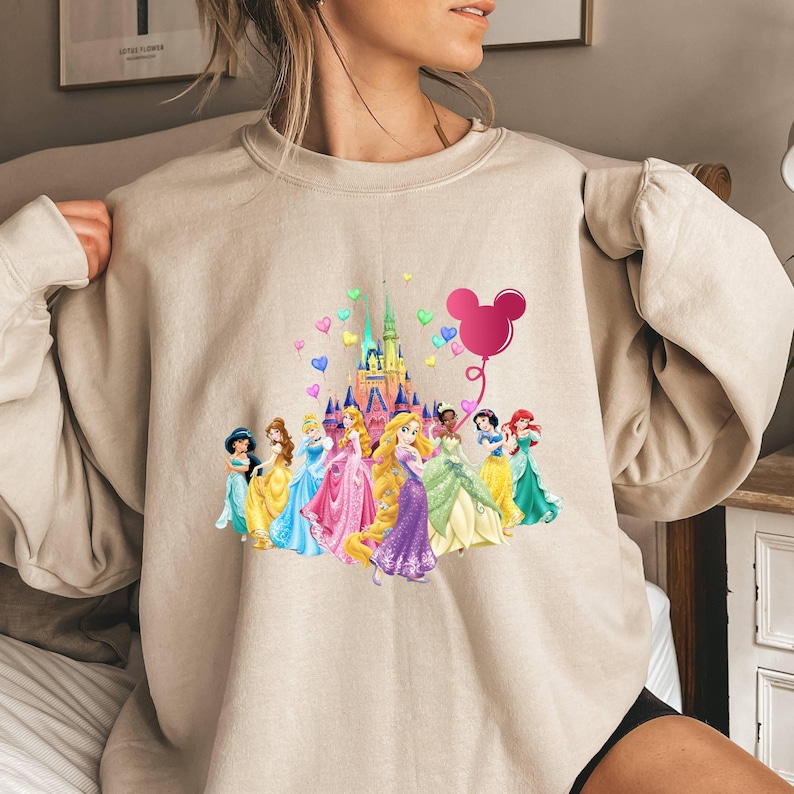 May include: A beige sweatshirt with a graphic print of Disney princesses and Cinderella's castle. The princesses are holding hands and walking towards the castle. There is a pink Mickey Mouse balloon in the air. The sweatshirt has a crew neck and long sleeves.