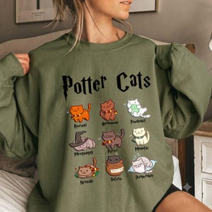 May include: An olive green crewneck sweatshirt featuring the text "Potter Cats" in black. Below the text are nine illustrated cats with names including Rawnald, Hermeownie, Pawdamort, Mewganagall, Puttier, Meowfay, Nyanville, Sefurus, and Dumpurr dore.