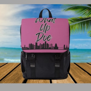 May include: Black and pink backpack with the text "What Up Doe" and a city skyline graphic. The backpack has a top handle, adjustable straps, and two front buckle closures. The bag is sitting on a wooden surface.