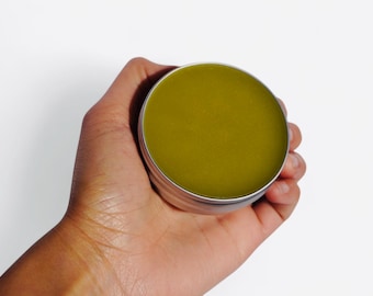 May include: A small round tin container filled with a green balm or ointment. The container is made of metal and has a silver lid.