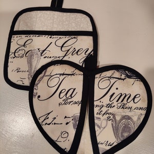 May include: Two black and white pot holders with a vintage design. One pot holder is square and says "Eat Grey", the other is heart shaped and says "Tea Time".