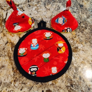 May include: A set of three red and black pot holders with a Japanese monkey design. The pot holders are round and have a black trim. The monkeys are wearing different outfits and hats.