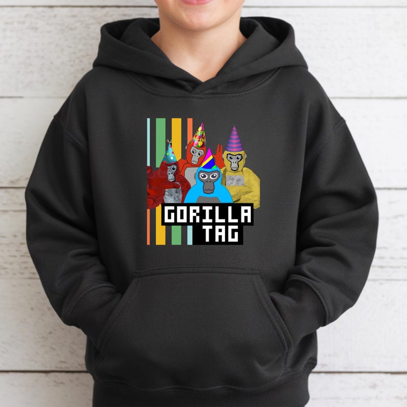 Puede incluir: Sudadera con capucha negra con un gr&aacute;fico de cuatro gorilas de dibujos animados con sombreros de fiesta. El texto "GORILLA TAG" se muestra en una fuente pixelada debajo de los gorilas, con rayas verticales en el lado izquierdo.