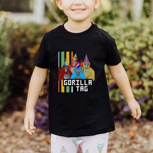Puede incluir: Camiseta negra con un gr&aacute;fico de cinco gorilas de dibujos animados con sombreros de fiesta. El texto "GORILLA TAG" se muestra debajo de los gorilas en una fuente pixelada, con rayas verticales de colores a la izquierda.