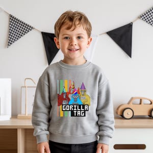 Puede incluir: Una sudadera gris con un gr&aacute;fico de tres gorilas de dibujos animados con sombreros de fiesta. El texto "GORILLA TAG" se muestra en una fuente pixelada. La sudadera la lleva un ni&ntilde;o peque&ntilde;o.