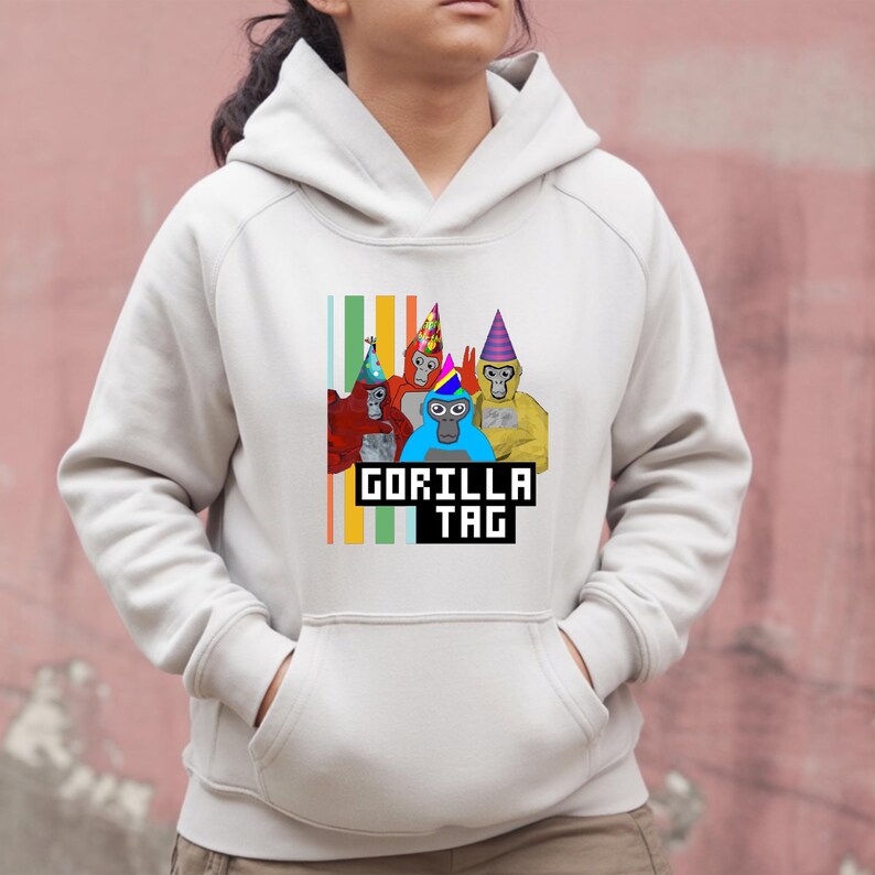 Puede incluir: Una sudadera con capucha blanca con un gr&aacute;fico colorido de cuatro gorilas de dibujos animados con sombreros de fiesta. El texto "GORILLA TAG" se muestra en una fuente pixelada debajo de los gorilas. La sudadera tiene un bolsillo de canguro.