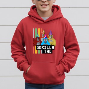 Puede incluir: Sudadera con capucha roja con un gr&aacute;fico de cuatro gorilas de dibujos animados con sombreros de fiesta. El texto "GORILLA TAG" se muestra en una fuente pixelada debajo de los gorilas. Rayas verticales de varios colores est&aacute;n en el lado izquierdo del gr&aacute;fico.