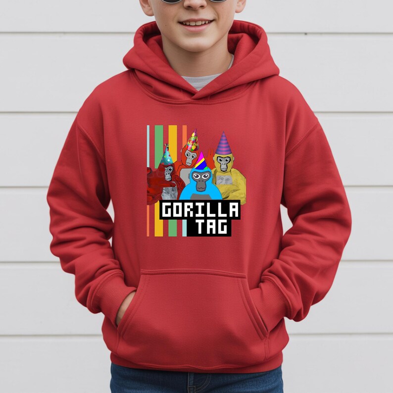 Puede incluir: Sudadera con capucha roja con un gr&aacute;fico de cuatro gorilas de dibujos animados con sombreros de fiesta. El texto "GORILLA TAG" se muestra en una fuente pixelada debajo de los gorilas. Rayas verticales de varios colores est&aacute;n en el lado izquierdo del gr&aacute;fico.