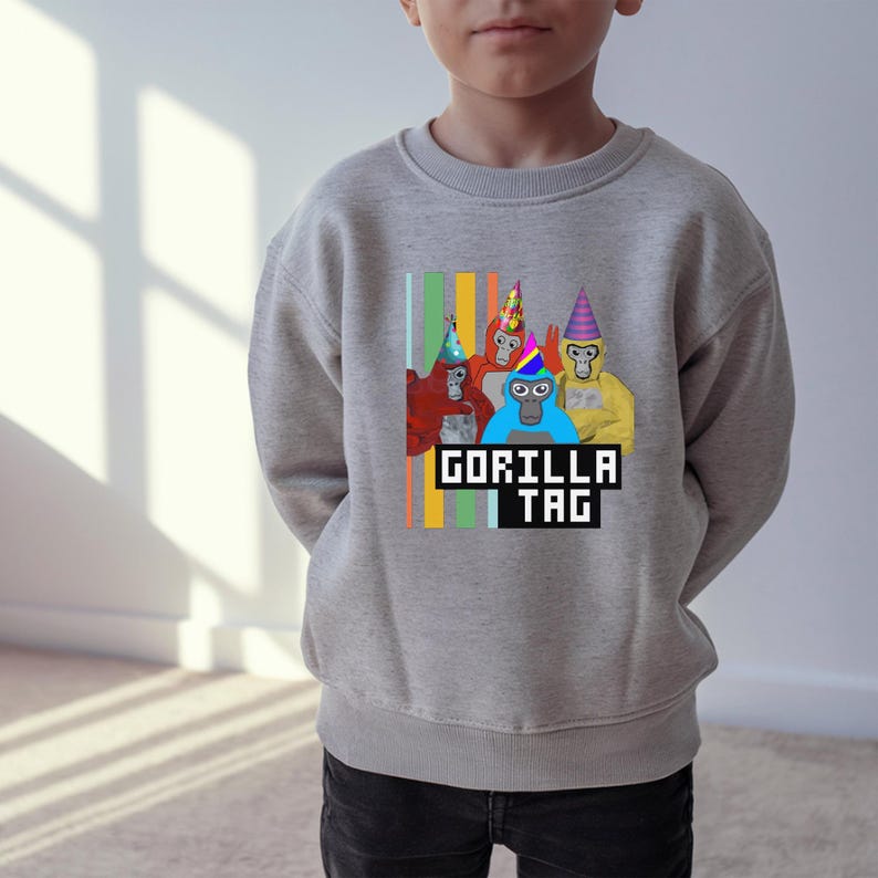Puede incluir: Una sudadera gris claro con un gr&aacute;fico de cuatro gorilas de dibujos animados con sombreros de fiesta. El texto "GORILLA TAG" se muestra en una fuente pixelada debajo de los gorilas, con rayas verticales de colores a la izquierda.
