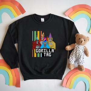 Puede incluir: Sudadera negra con un gr&aacute;fico de cinco gorilas de dibujos animados con sombreros de fiesta. El texto "GORILLA TAG" se muestra en una fuente pixelada debajo de los gorilas. Rayas verticales en verde, amarillo y naranja a la izquierda.