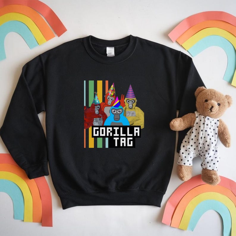 Puede incluir: Sudadera negra con un gr&aacute;fico de cinco gorilas de dibujos animados con sombreros de fiesta. El texto "GORILLA TAG" se muestra en una fuente pixelada debajo de los gorilas. Rayas verticales en verde, amarillo y naranja a la izquierda.