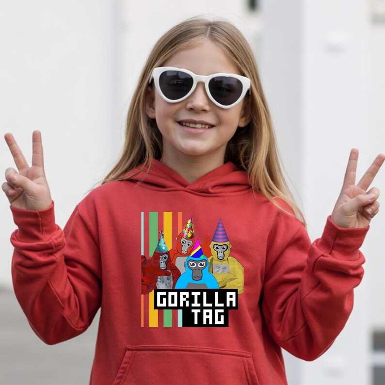 Puede incluir: Una sudadera con capucha roja con un gr&aacute;fico de cuatro gorilas de dibujos animados con sombreros de fiesta. El texto "GORILLA TAG" se muestra en una fuente pixelada. La persona de la imagen lleva gafas de sol blancas y hace el signo de la paz.