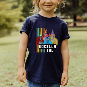 Puede incluir: Camiseta azul marino con un gr&aacute;fico de cuatro gorilas de dibujos animados con sombreros de fiesta. El texto "GORILLA TAG" se muestra en una fuente pixelada. Rayas verticales de varios colores est&aacute;n en el lado izquierdo del gr&aacute;fico.