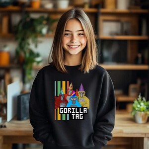 Puede incluir: Sudadera negra con un gr&aacute;fico de cuatro gorilas de dibujos animados con sombreros de fiesta. El texto "GORILLA TAG" se muestra en una fuente pixelada debajo de los gorilas. La sudadera la lleva una persona.