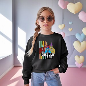 Puede incluir: Sudadera negra con un gr&aacute;fico de cuatro gorilas de dibujos animados con sombreros de fiesta. El texto "GORILLA TAG" se muestra en una fuente pixelada debajo de los gorilas. El dise&ntilde;o est&aacute; sobre un fondo de rayas verticales de varios colores.