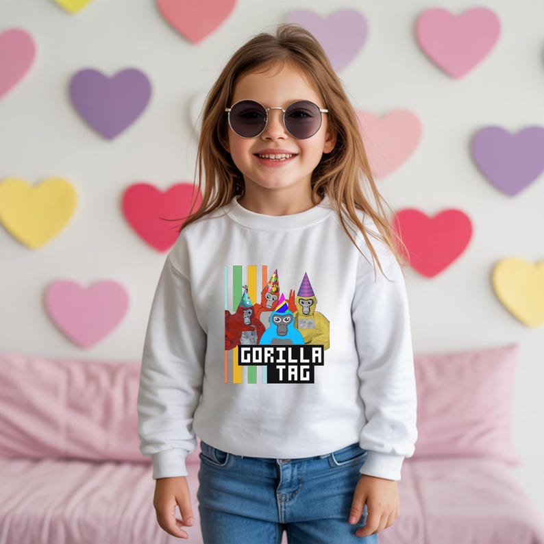Puede incluir: Sudadera blanca con un gr&aacute;fico de gorilas con sombreros de fiesta y el texto "GORILLA TAG". El dise&ntilde;o incluye rayas verticales de varios colores. La sudadera la lleva un ni&ntilde;o.