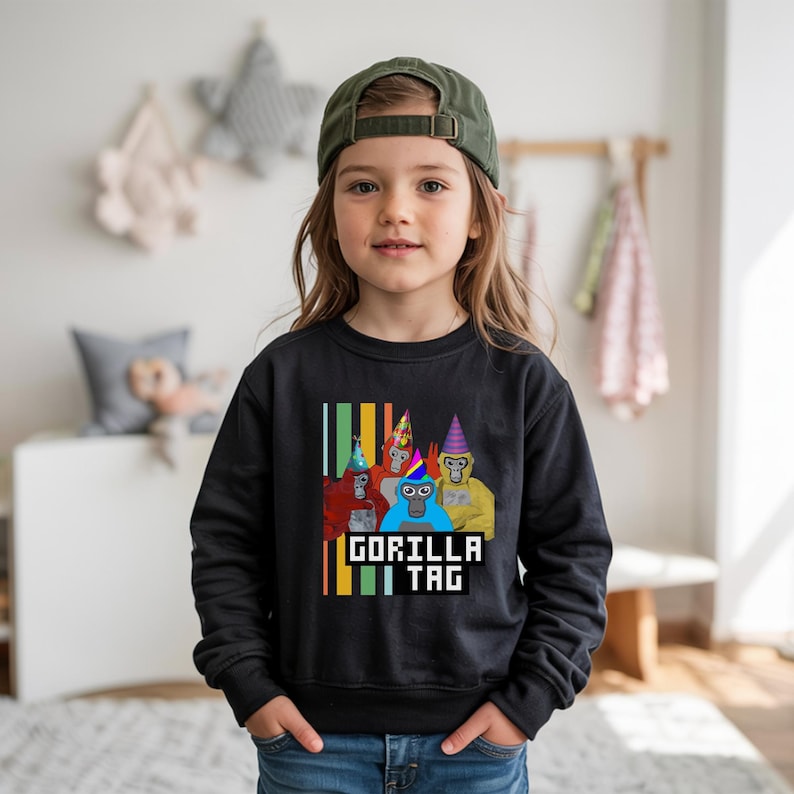 Puede incluir: Sudadera negra con un gr&aacute;fico de gorilas de dibujos animados con sombreros de fiesta. El texto "GORILLA TAG" se muestra en una fuente pixelada. La sudadera la lleva un ni&ntilde;o peque&ntilde;o.