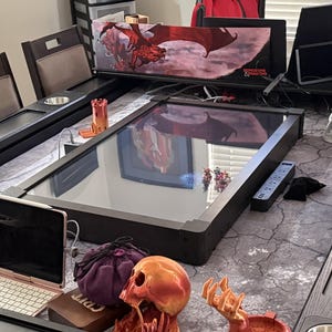 May include: A rectangular gaming table featuring a central digital screen, a Dungeons & Dragons screen, a tablet with a keyboard, a purple drawstring bag, a skull-shaped object, a hand-shaped tray, and a small tower figurine on a gray mat.