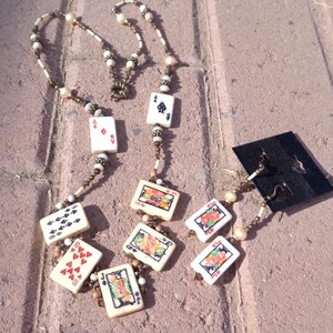 May include: A necklace and earrings set featuring playing card pendants. The necklace has a beaded chain with playing card charms, including the Ace of Spades, Ace of Diamonds, and others. The earrings have similar playing card charms. The set is displayed on a brick surface.