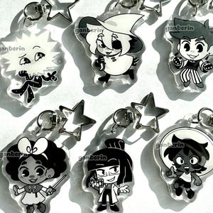 May include: A set of six clear acrylic keychains featuring black and white cartoon character illustrations. Each keychain includes a silver-toned star-shaped clasp. The characters include a sun-like figure, a witch, a person with striped pants, a figure with a bow, a person in a lab coat, and a character in a circular frame. Each piece is marked with the text ganberin.