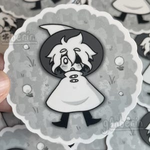 May include: A grayscale sticker depicting a cartoon character with white hair, a hat, and a cloak. The character is set against a grey background with a scalloped edge, with small white circles and grass details.