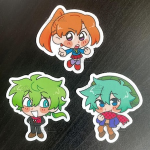May include: Three anime-style chibi character stickers. One has orange hair, a blue jacket, and a red skirt. The other two have green hair, one with a black suit and red scarf, the other with a green hat and a red scarf.