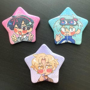 May include: Three star-shaped badges with glittery surfaces. Each badge features a cartoon character with different hair colours and outfits. The badges are pink, blue, and purple, with the characters in various poses.