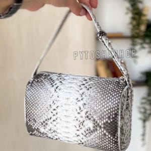 Peut inclure: Un petit sac à main gris argenté avec un motif en peau de serpent. Le sac a une courte bandoulière et une fermeture à glissière.