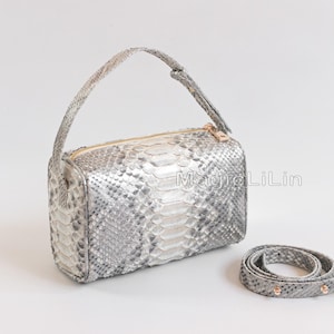 Könnte beinhalten: Eine rechteckige Handtasche aus silber- und weißfarbenem Schlangenleder mit goldenem Reißverschluss und Griff. Ein passender Riemen ist in der Nähe aufgerollt. Auf der Tasche steht der Text "Magic LiLin".
