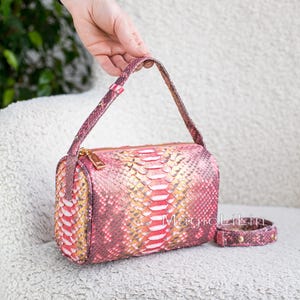 May include: A pink and gold snakeskin-patterned handbag with a matching bracelet. The rectangular bag has a top handle and a gold zip. The bracelet has gold studs. The bag and bracelet are on a white chair.