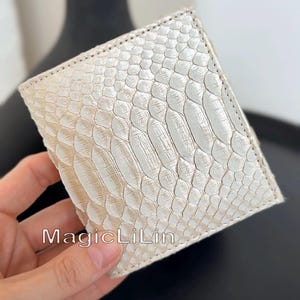 May include: A square-shaped wallet featuring a pearlescent white snakeskin texture with a visible scale pattern. The wallet has stitched edges and the text MagicLiLin is printed in a clear font on the lower portion of the front surface.