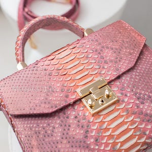 May include: A pink handbag with a snakeskin pattern, featuring a top handle and a detachable shoulder strap. The bag has a gold-colored clasp and the text "MagicLilin" is visible on the front.