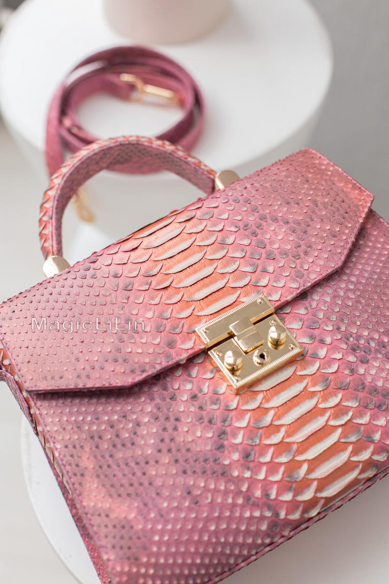 May include: A pink handbag with a snakeskin pattern, featuring a top handle and a detachable shoulder strap. The bag has a gold-colored clasp and the text "MagicLilin" is visible on the front.