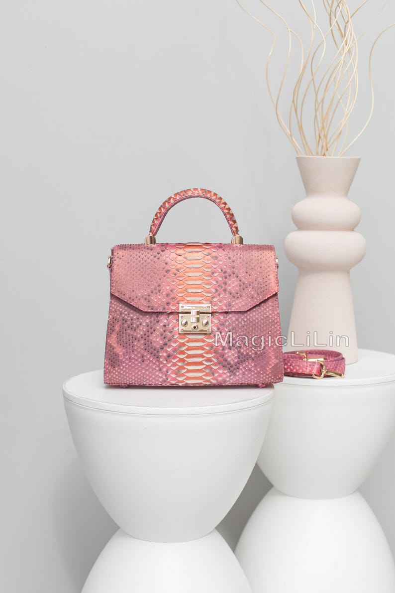 May include: Pink and orange snakeskin handbag with a top handle and a gold clasp. A matching strap is next to the bag. The bag is on a white table. The text "MagicLiLin" is on the bag.