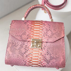May include: A pink handbag with a snakeskin pattern, featuring a top handle and a detachable shoulder strap. The bag has a gold-colored clasp and hardware. The snakeskin pattern transitions from pink to orange in the center.