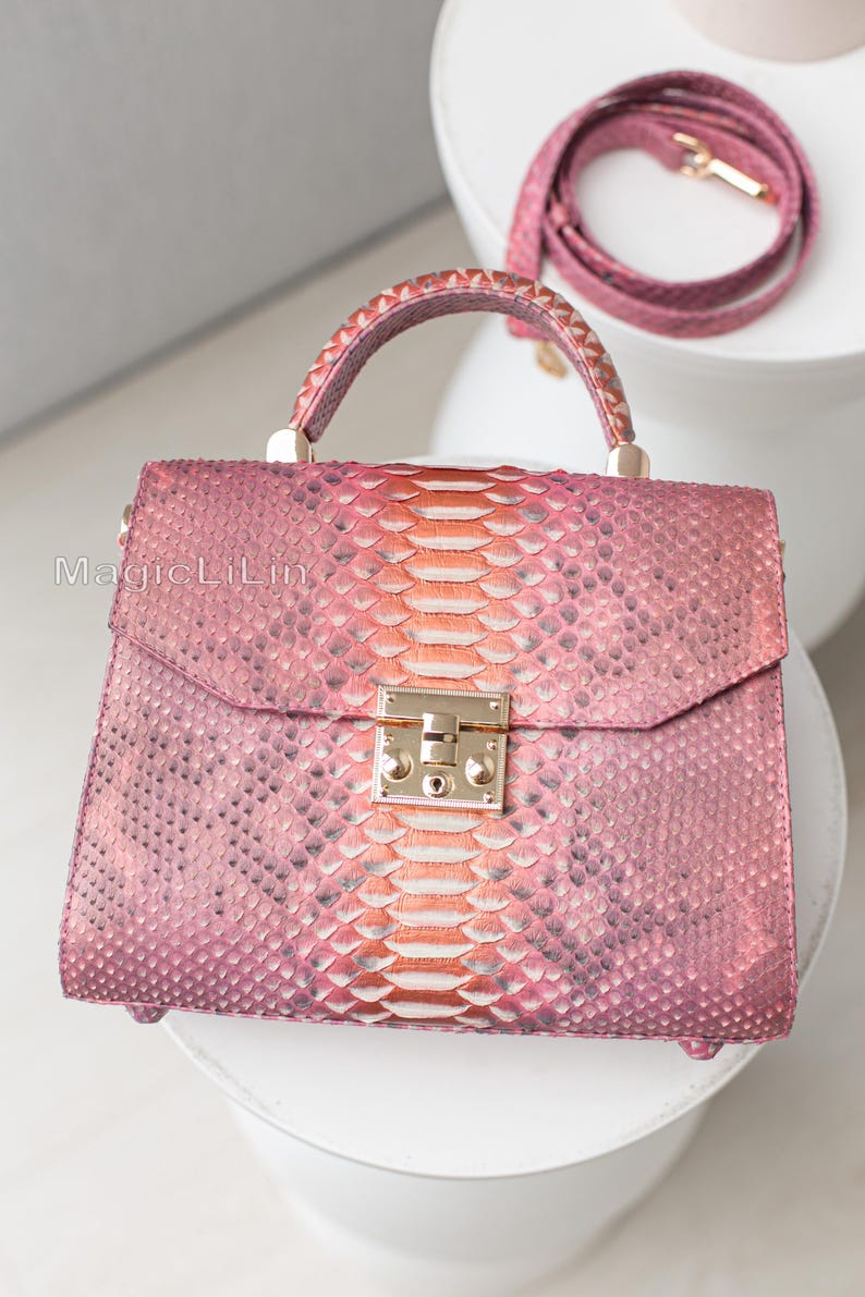 May include: A pink handbag with a snakeskin pattern, featuring a top handle and a detachable shoulder strap. The bag has a gold-colored clasp and hardware. The snakeskin pattern transitions from pink to orange in the center.