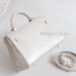 May include: A light gray handbag with a textured snakeskin pattern. The bag has a top handle and a detachable shoulder strap. The text "MagicLiLin" is printed on the side of the bag. The hardware is gold-colored.