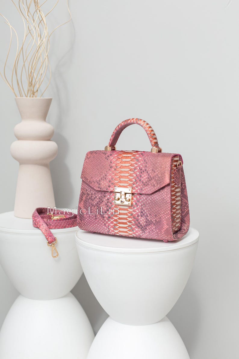 May include: A pink snakeskin handbag with a top handle and a detachable shoulder strap. The bag features a gold-tone clasp and hardware. The bag is displayed on a white pedestal.