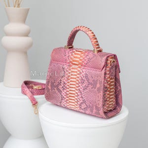 May include: Pink handbag with a snakeskin pattern, featuring a top handle and a detachable shoulder strap. The bag has gold-colored hardware and is displayed on a white surface. The text "MagicLilin" is visible on the side of the bag.