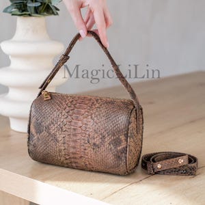 Könnte beinhalten: Eine braune Schlangenleder-Handtasche mit einem goldenen Reißverschluss und einem passenden Riemen. Die Tasche liegt auf einer hellbraunen Holzoberfläche.