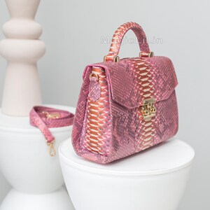 May include: A pink snakeskin handbag with a top handle and a detachable shoulder strap. The bag features a gold-colored clasp and hardware. The text "MagicLiLin" is visible on the bag. The bag is displayed on a white surface.