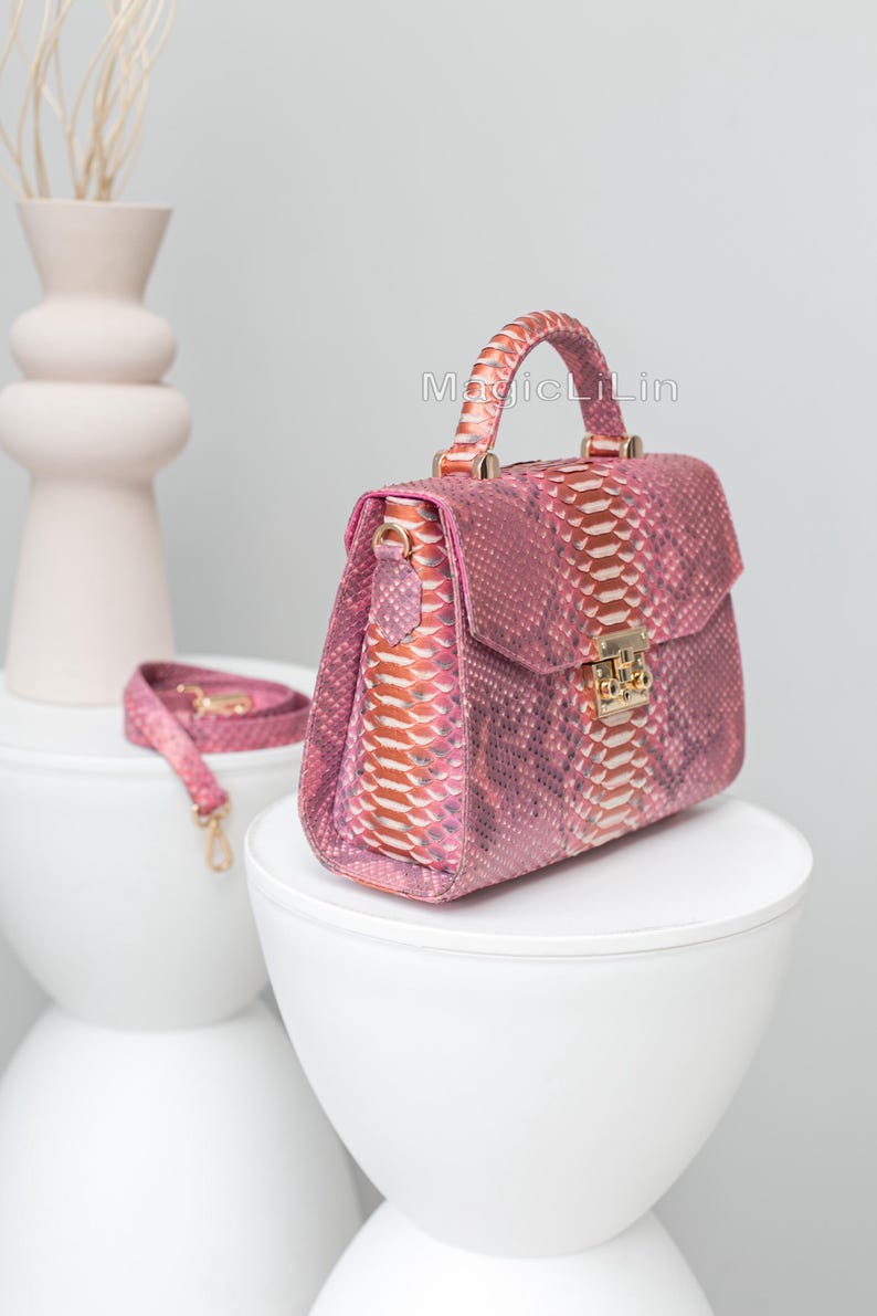 May include: A pink snakeskin handbag with a top handle and a detachable shoulder strap. The bag features a gold-colored clasp and hardware. The text "MagicLiLin" is visible on the bag. The bag is displayed on a white surface.