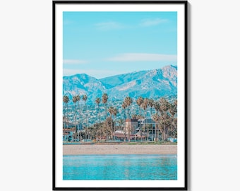 May include: A framed landscape photograph featuring a turquoise ocean in the foreground, a sandy beach, a row of tall palm trees, and residential buildings nestled at the base of a large, rugged mountain range under a clear light blue sky.