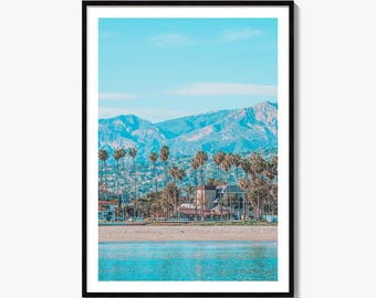 May include: A framed wall art print featuring a coastal landscape with a turquoise ocean in the foreground, a sandy beach, a row of tall palm trees, and a backdrop of rugged blue mountains under a clear, bright sky during the daytime.