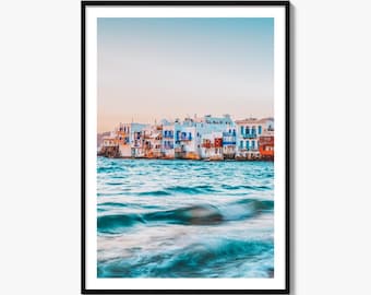May include: A framed wall art print featuring a coastal scene with white buildings with blue accents situated along the water's edge. The foreground shows turquoise ocean waves with a soft motion blur effect under a clear, pale sky at sunset.