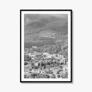 Könnte beinhalten: Ein schwarz-weiß gerahmter Druck mit einer Luftaufnahme einer Stadt, die am Fuße eines großen, bewaldeten Gebirgszuges liegt. Die Landschaft umfasst verschiedene Gebäude, Straßen und dichte Bäume unter einem bewölkten Himmel.