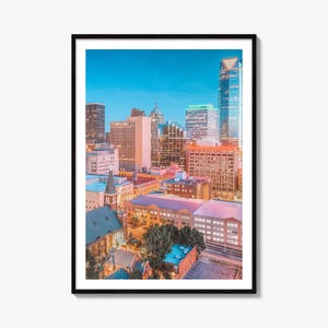 May include: A framed vertical wall art print featuring a high-angle cityscape of Dallas, Texas, at twilight. The scene displays various skyscrapers, historic buildings, and urban architecture illuminated by warm orange and cool blue light tones.